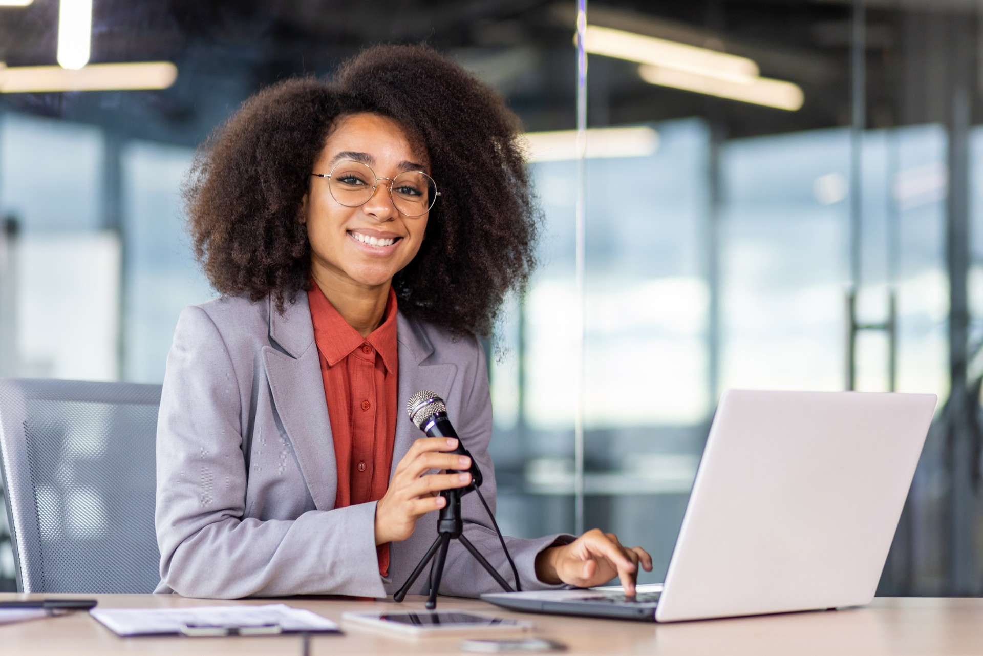 Mujer negra satisfecha con gafas que arregla el micrófono profesional conectado a una computadora portátil en el escritorio. Podcaster femenina preparándose para grabar audio de alta calidad en un estudio equipado